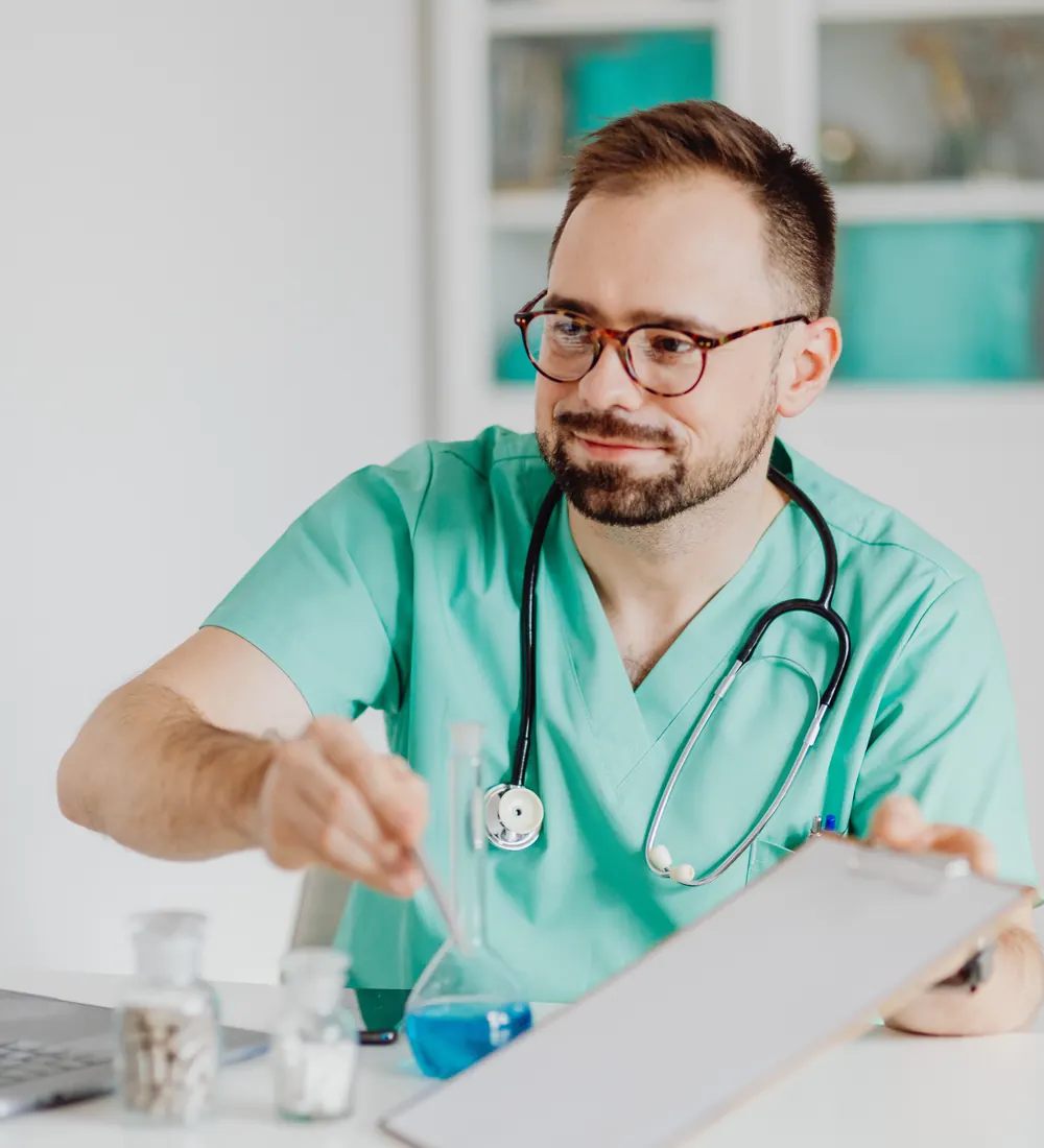 Healthcare provider in scrubs with a stethoscope reviewing notes on a clipboard at a desk