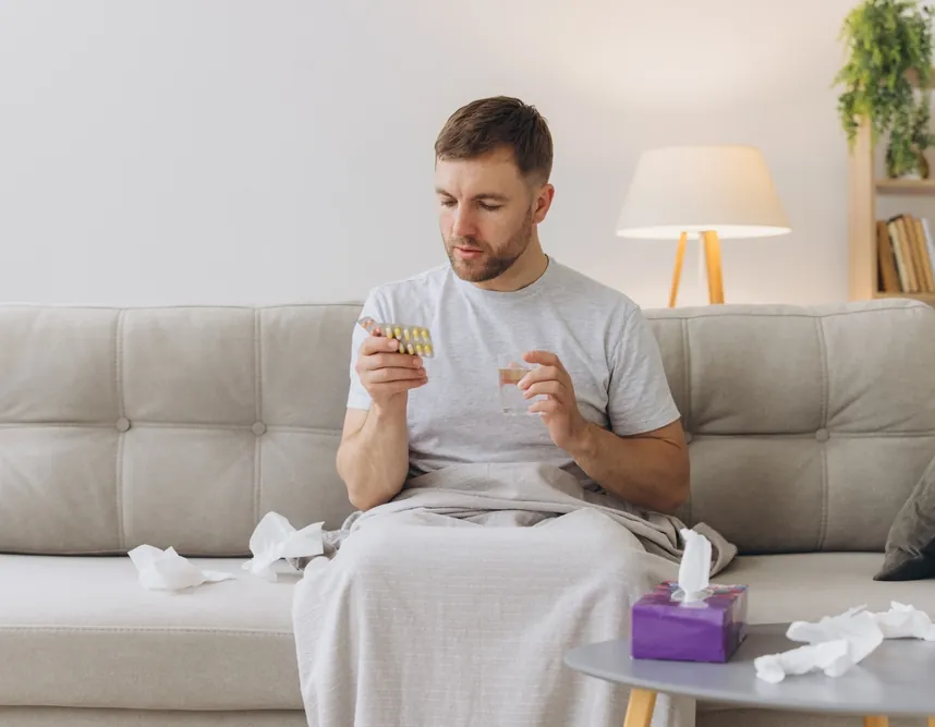 Man sitting on a sofa holding a blister pack of tablets and a glass of water, representing testosterone therapy for men in St. Petersburg, FL